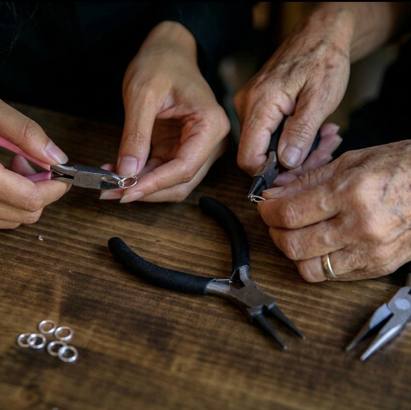 Hands working with jewelry on a wooden table with pliers and rings.