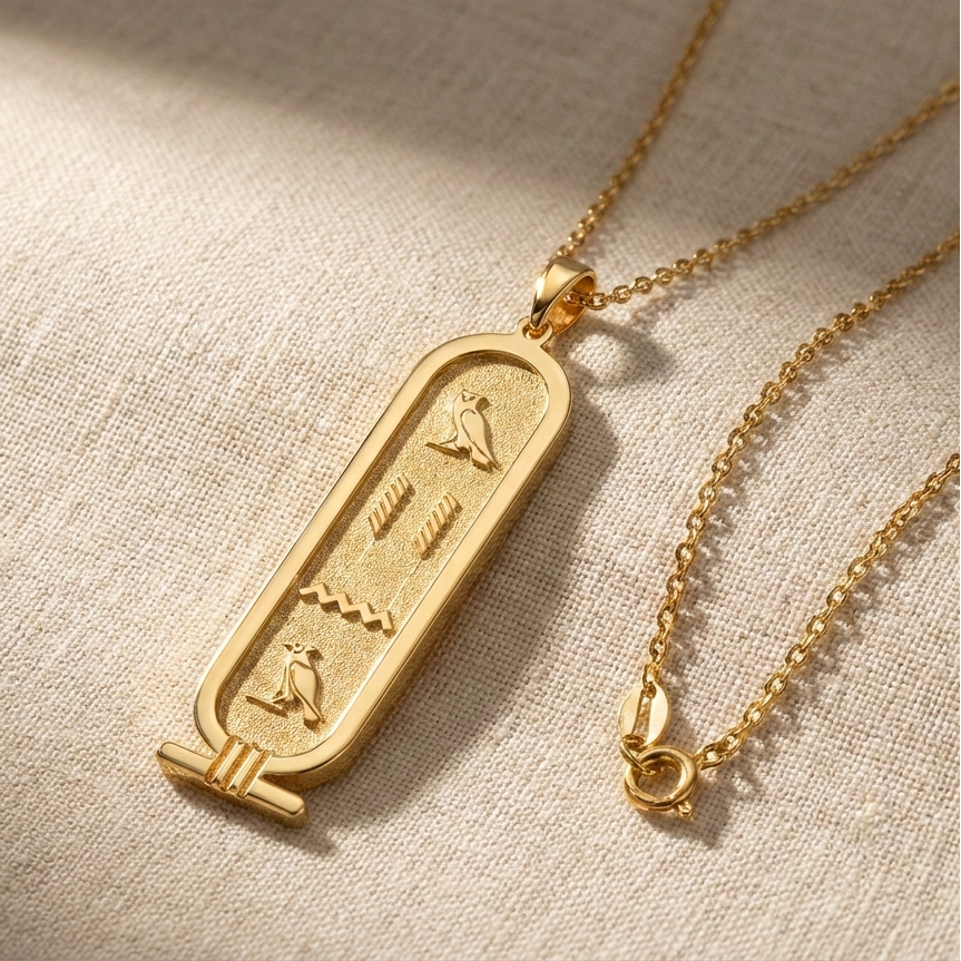Gold necklace with an oval pendant featuring hieroglyphics on a beige fabric background