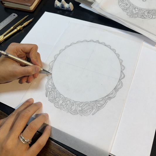 Person drawing intricate jewelry on paper with a pen, surrounded by stationery items.