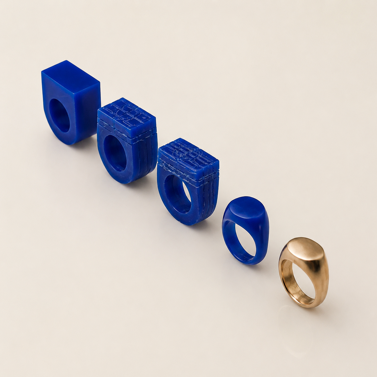 Four sculptural rings arranged diagonally on a warm off-white background, including three in glossy cobalt blue with carved text details and one in polished gold.