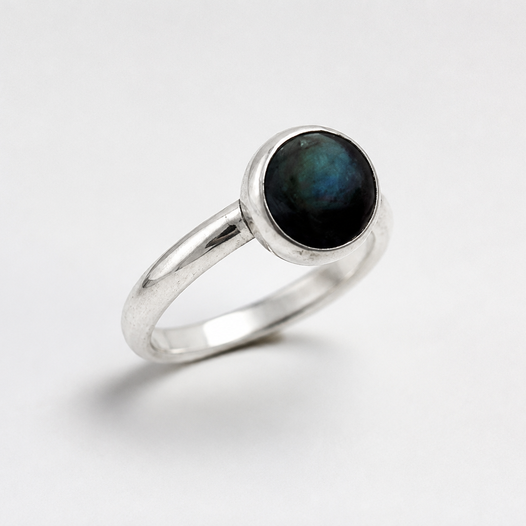 A silver ring featuring a round bezel-set dark iridescent stone, photographed on a clean white background.