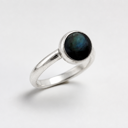 A silver ring featuring a round bezel-set dark iridescent stone, photographed on a clean white background.